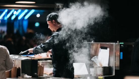Vue d'un cuisinier de dos, il est en tenue noire, un nuage de vapeur monte derrière lui